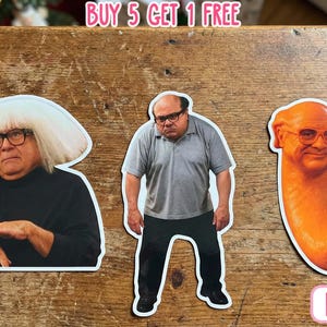 May include: Three die-cut stickers featuring images of a man with glasses. One sticker shows the man with a white wig, another shows him in a gray shirt, and the third is a caricature of his face on a hot dog. The stickers are on a wooden surface.