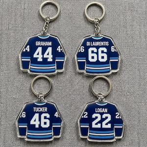 May include: Four hockey jersey keyrings. Each keyring is navy blue with white and light blue stripes, displaying a player's name and number. The names are Graham, Di Laurentis, Tucker, and Logan. The numbers are 44, 66, 46, and 22.