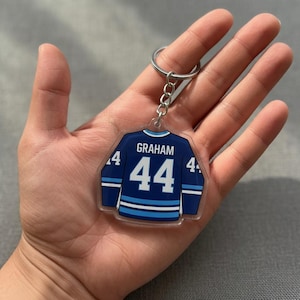 May include: A clear acrylic hockey jersey-shaped keychain. The navy blue jersey features light blue and white stripes, the number 44, and the name "GRAHAM". It is attached to a silver metal ring.