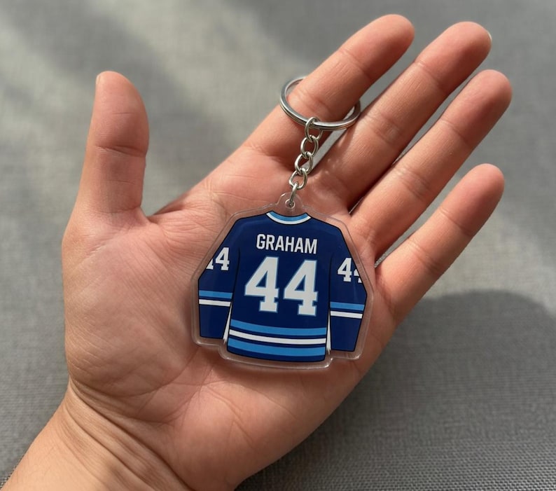 May include: A clear acrylic hockey jersey-shaped keychain. The navy blue jersey features light blue and white stripes, the number 44, and the name "GRAHAM". It is attached to a silver metal ring.