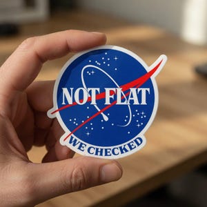 Puede incluir: Una pegatina redonda con fondo azul y estrellas blancas, con el texto "NOT FLAT" y "WE CHECKED" en blanco. Una línea roja cruza el centro del diseño, similar a un logotipo de la NASA.