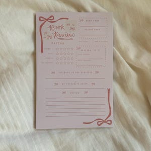 May include: A pale pink book review card with bow illustrations. The card features sections for rating, book details, and a quote. The text "Book Review" is in cursive. The card is designed for book lovers.