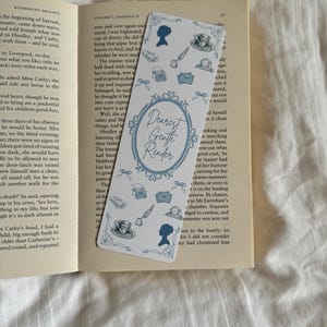 May include: A rectangular white bookmark featuring blue illustrations of silhouettes, teacups, envelopes, and quills. The centre displays an ornate blue oval frame containing the text Dearest Gentle Reader in a cursive script font.