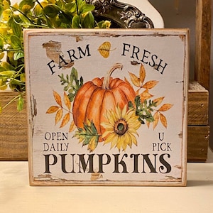 May include: A rustic wooden sign with the text "FARM FRESH PUMPKINS" in black lettering. The sign features a watercolor illustration of an orange pumpkin, sunflower, and autumn leaves. The sign is square and has a distressed, vintage look.