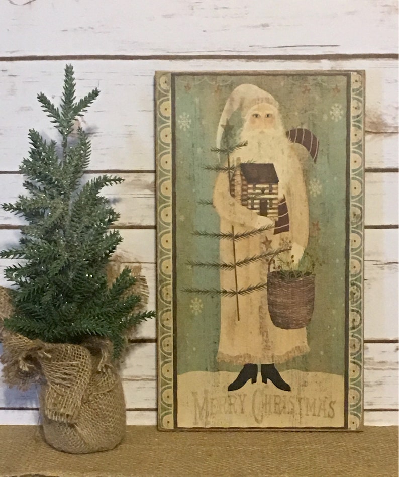 May include: A vintage Christmas wooden sign with an illustration of Santa Claus holding a tree and basket. The sign has a distressed finish and reads "Merry Christmas". A small evergreen tree in a burlap pot is positioned to the left.