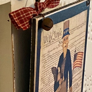 May include: A wooden sign with a blue border and a red, white, and blue illustration of Uncle Sam holding a flag and an eagle. The sign says "We the People" and is decorated with a red and white plaid ribbon and a bell.