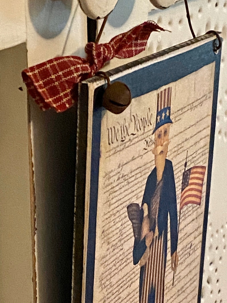 May include: A wooden sign with a blue border and a red, white, and blue illustration of Uncle Sam holding a flag and an eagle. The sign says "We the People" and is decorated with a red and white plaid ribbon and a bell.