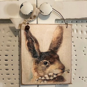 May include: A vintage-style wooden sign with a brown rabbit illustration. The rabbit is holding a sprig of white flowers in its mouth. The sign has a distressed white background and is hung with a white ribbon and two white knobs.