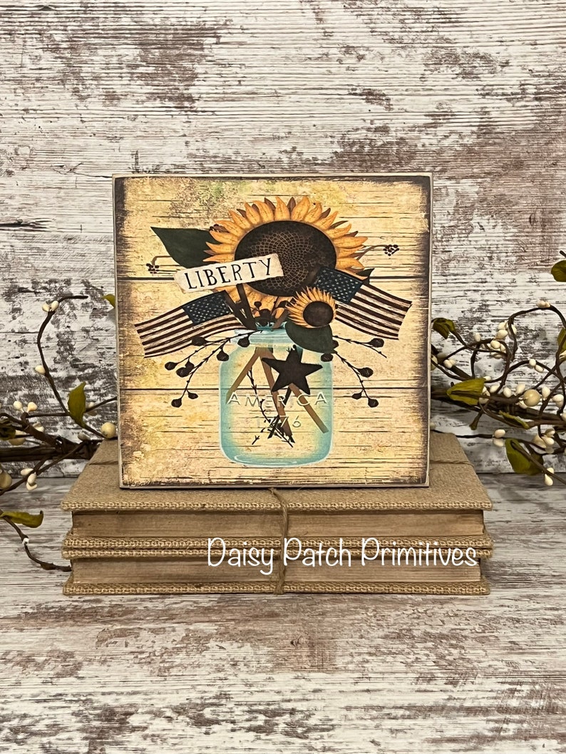 May include: A wooden sign with a rustic background featuring a mason jar filled with sunflowers and American flags. The jar has the words "America 1776" on it. The sign also has a small sign that says "Liberty".