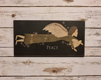 May include: A rectangular wooden sign with a black background features a stylized angel in flight, holding a dove. The angel has tan wings and a green dress. The word "PEACE" is written in white. Snowflakes are scattered around the angel.