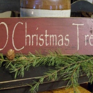 Puede incluir: Un letrero de madera rústico y rectangular con las palabras "O Christmas Tree" en letras blancas. El letrero está pintado de color rojo oscuro y está decorado con una piña y una guirnalda de hojas perennes. El letrero está colocado sobre una superficie negra.