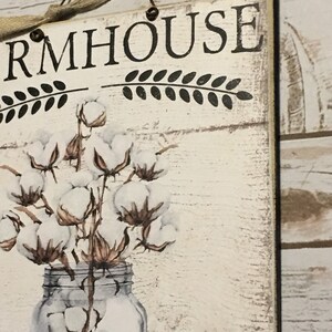 May include: A white wooden sign with the text "FARMHOUSE" in black lettering. The sign features a decorative design of cotton branches in a clear glass jar.