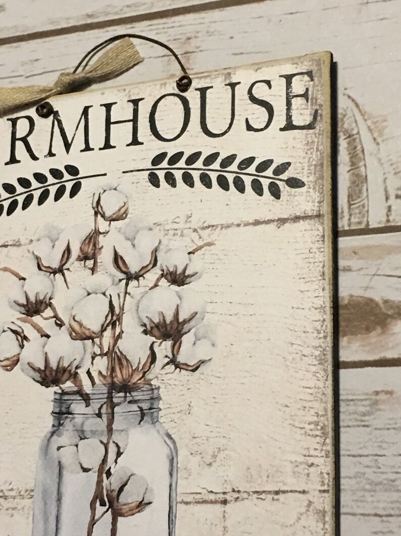 May include: A white wooden sign with the text "FARMHOUSE" in black lettering. The sign features a decorative design of cotton branches in a clear glass jar.