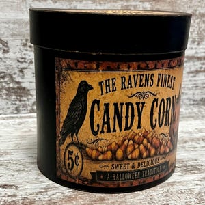 May include: A round black container with a vintage-style label featuring a black raven, a pile of candy corn, and the text THE RAVENS FINEST CANDY CORN, 5 cents, SWEET & DELICIOUS, and A HALLOWEEN TRADITION printed in black on a tan background.