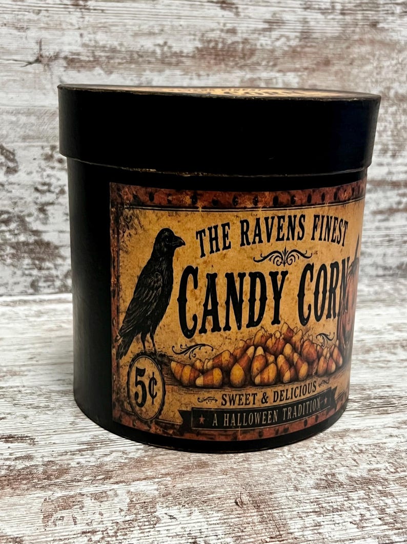 May include: A round black container with a vintage-style label featuring a black raven, a pile of candy corn, and the text THE RAVENS FINEST CANDY CORN, 5 cents, SWEET & DELICIOUS, and A HALLOWEEN TRADITION printed in black on a tan background.