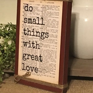 May include: A rustic wooden sign with a red frame and twine accents. The sign features a dictionary page background with the text "do small things with great love" printed in black.