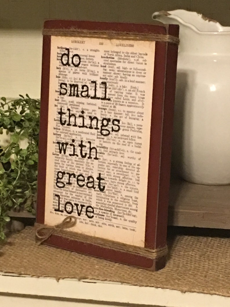 May include: A rustic wooden sign with a red frame and twine accents. The sign features a dictionary page background with the text "do small things with great love" printed in black.