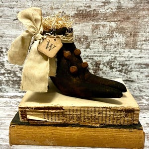 May include: A dark brown fabric boot ornament with a pointed toe, adorned with small rusted metal bells, a cream-colored fabric bow, and a small tan tag featuring the letter W. The boot is filled with straw and rests atop two stacked books.