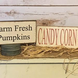May include: Two rustic wooden signs with text. One reads "Farm Fresh Pumpkins" in black on a cream background, mounted on a metal base. The other says "CANDY CORN" in red. Both are set on a bed of straw.