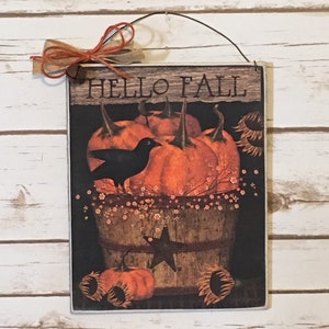 May include: Wooden sign with the words "HELLO FALL" above a fall-themed illustration. The illustration features pumpkins, sunflowers, and a black crow in a wooden bucket. A small bow is attached to the top.