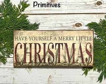 May include: A rectangular wooden sign with the text "HAVE YOURSELF A MERRY LITTLE CHRISTMAS" in burgundy letters. The sign is decorated with sprigs of greenery and set against a white wooden background. The words "Daisy Patch Primitives" are above the sign.