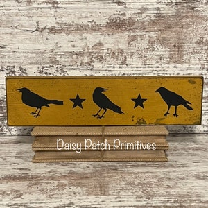 May include: A wooden sign with a yellow background and black silhouettes of three crows and two stars. The sign is sitting on a stack of burlap-covered books. The text "Daisy Patch Primitives" is visible on the books.
