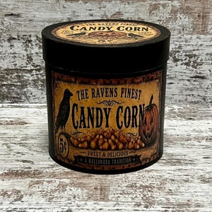 May include: A cylindrical black container with a vintage-style label featuring a black raven, a jack-o'-lantern, and a pile of candy corn. The text reads The Ravens Finest Candy Corn, Sweet & Delicious, A Halloween Tradition, and 5 cents.
