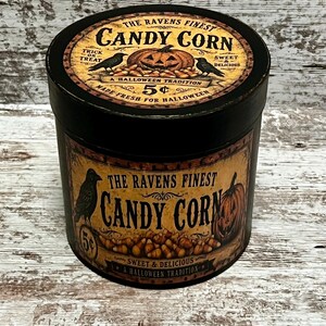 May include: A round black container with a vintage-style label featuring the text The Ravens Finest Candy Corn, a jack-o'-lantern, ravens, and candy corn illustrations. The label includes the phrases Trick or Treat, Sweet & Delicious, and 5 cents.