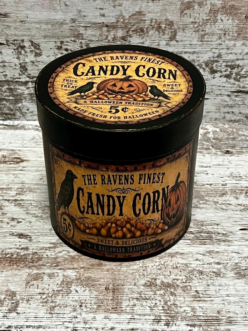 May include: A round black container with a vintage-style label featuring the text The Ravens Finest Candy Corn, a jack-o'-lantern, ravens, and candy corn illustrations. The label includes the phrases Trick or Treat, Sweet & Delicious, and 5 cents.
