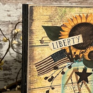 May include: A wooden sign with a distressed, rustic look. The sign features a mason jar filled with sunflowers, a small American flag, and a sign that reads "LIBERTY".