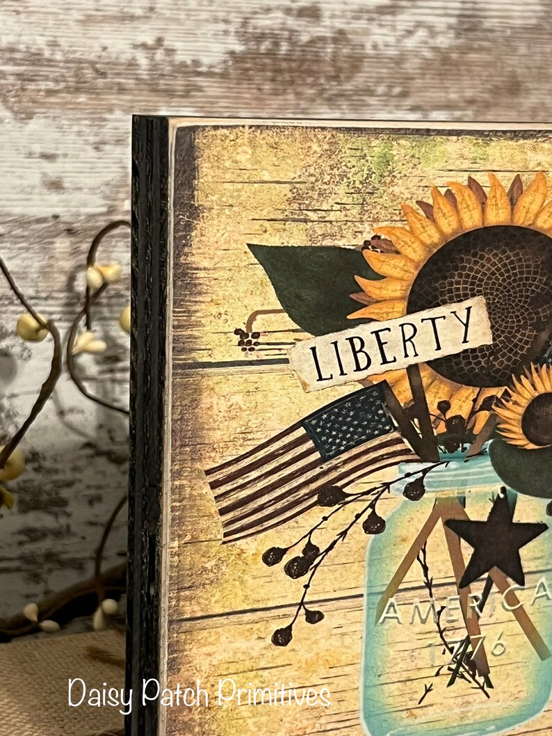May include: A wooden sign with a distressed, rustic look. The sign features a mason jar filled with sunflowers, a small American flag, and a sign that reads "LIBERTY".