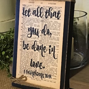 May include: A wooden sign with a black frame and twine. The sign features a vintage dictionary page with the text "Let all that you do, be done in love. 1 Corinthians 16:14" printed in black.