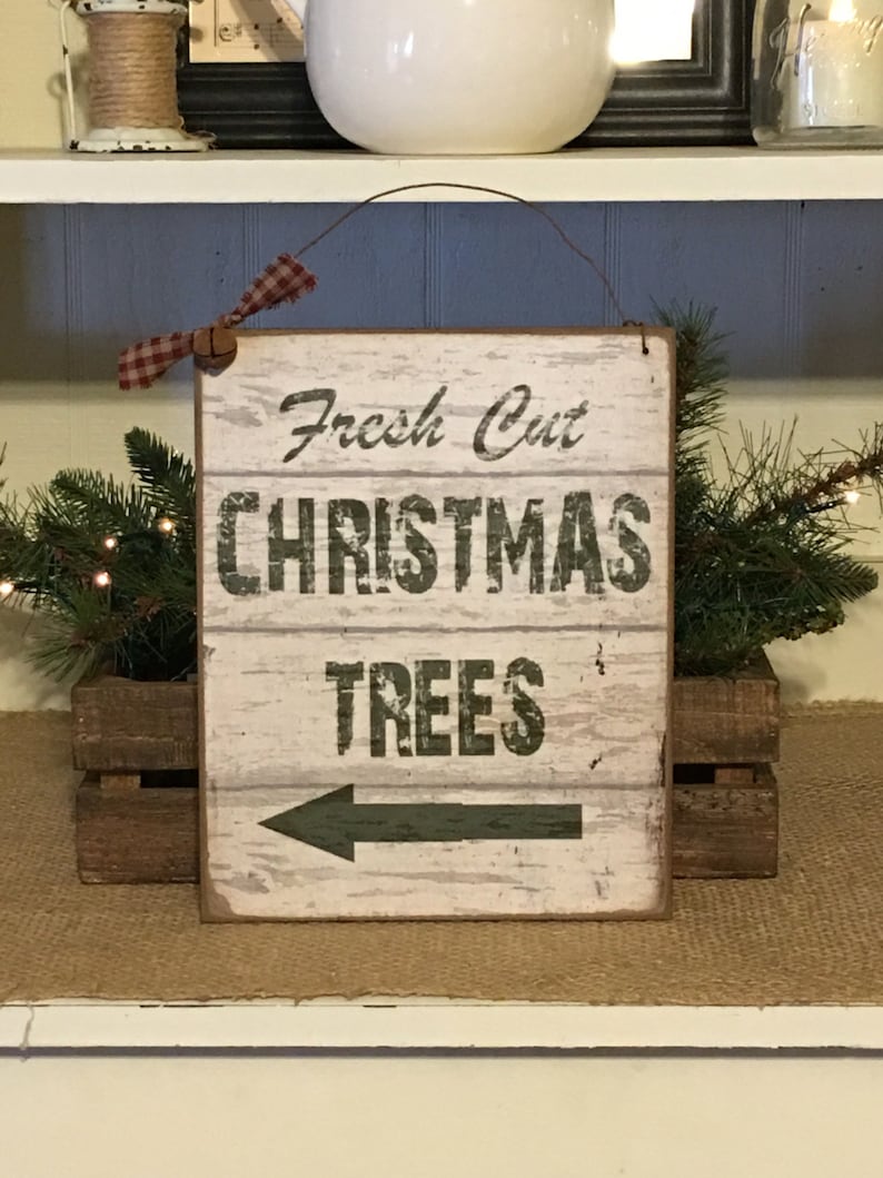 以下が含まれることがあります： 「Fresh Cut CHRISTMAS TREES」と緑色の文字で書かれた、木製の素朴な看板。左向きの矢印があります。赤と白のチェック柄のリボンで飾られ、緑に囲まれています。