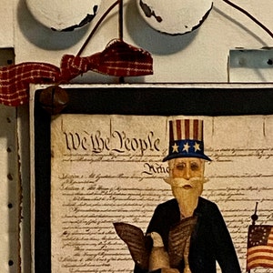 May include: A framed print of a cartoon Uncle Sam holding a bald eagle and an American flag. The print is framed in black and has a white background with the text "We the People" printed in black.