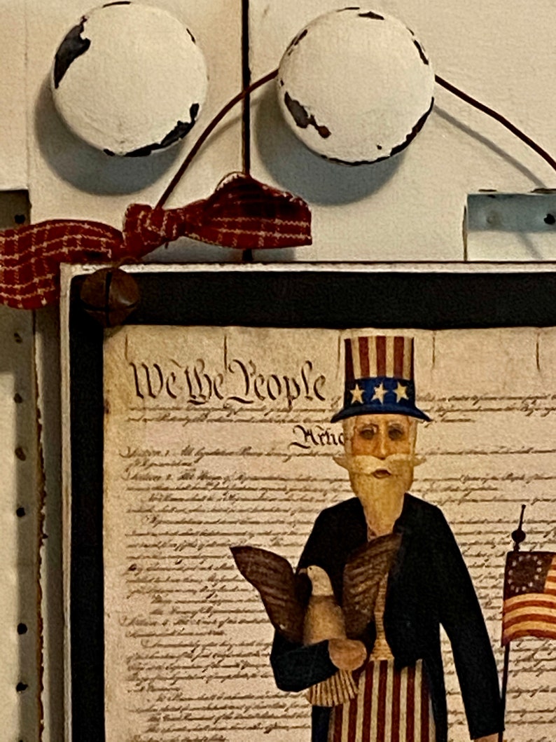 May include: A framed print of a cartoon Uncle Sam holding a bald eagle and an American flag. The print is framed in black and has a white background with the text "We the People" printed in black.