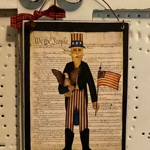 May include: A framed print of a folk art style illustration of Uncle Sam holding an eagle and an American flag. The print is on a black background and features the text "We the People" from the US Constitution.