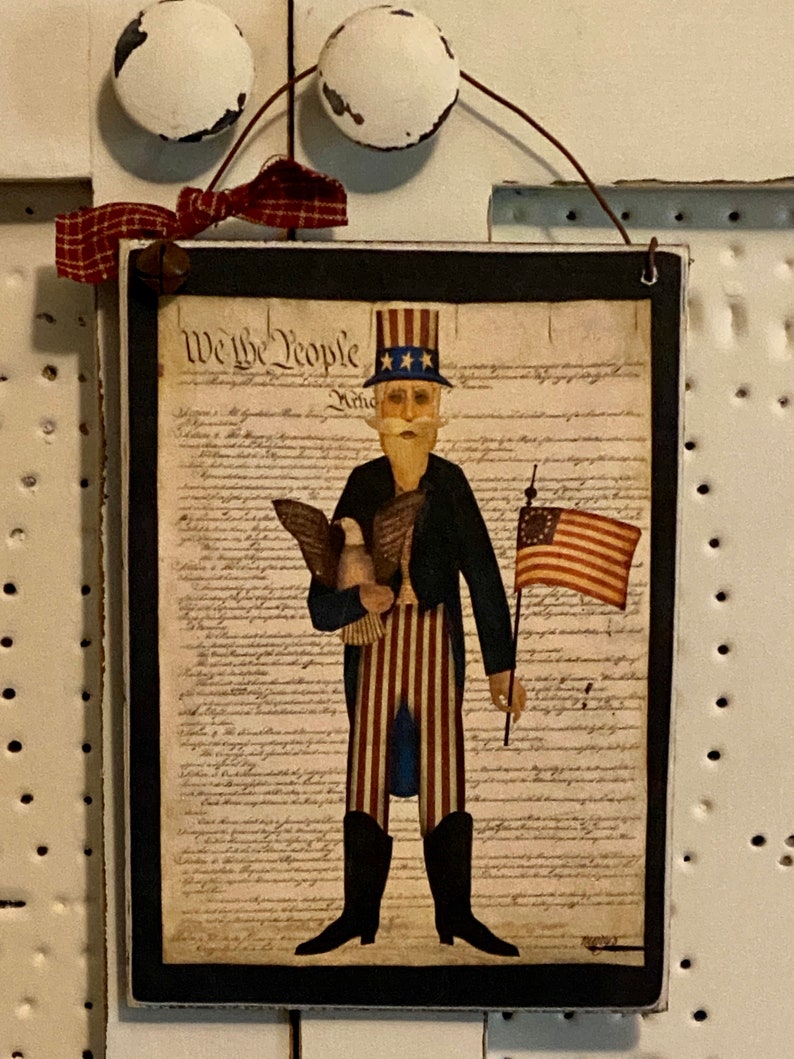 May include: A framed print of a folk art style illustration of Uncle Sam holding an eagle and an American flag. The print is on a black background and features the text "We the People" from the US Constitution.