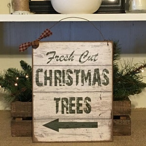 以下が含まれることがあります： 「Fresh Cut CHRISTMAS TREES」の文字と左向きの矢印が描かれた、素朴な木製の看板。 表面は使い古したような風合いで、赤と白のチェック柄のリボンが付いています。 木箱に入ったグリーンと共に飾られ、クリスマスの雰囲気を醸し出しています。
