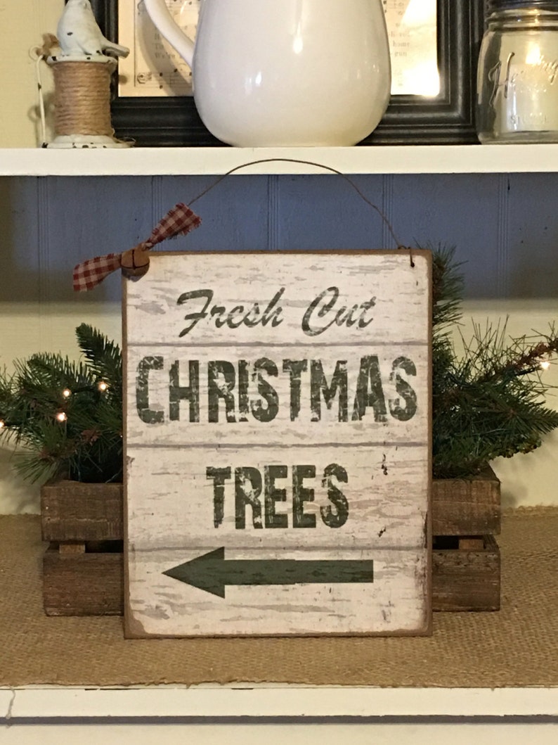 以下が含まれることがあります： 「Fresh Cut CHRISTMAS TREES」の文字と左向きの矢印が描かれた、素朴な木製の看板。 表面は使い古したような風合いで、赤と白のチェック柄のリボンが付いています。 木箱に入ったグリーンと共に飾られ、クリスマスの雰囲気を醸し出しています。