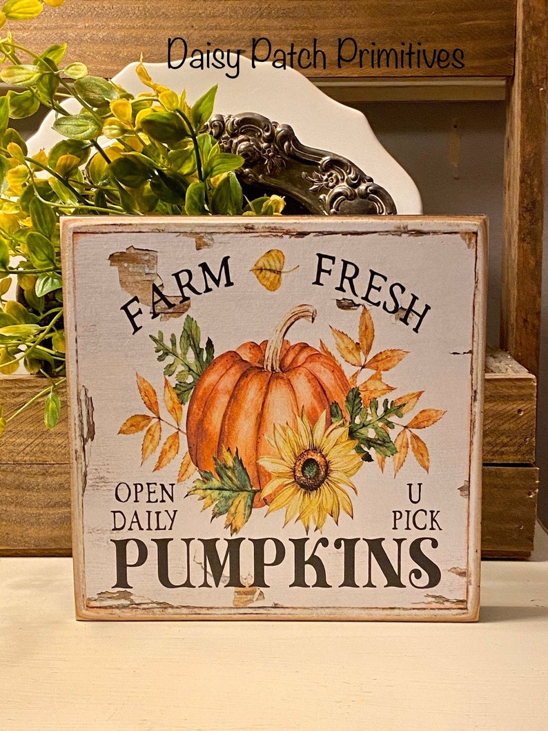 May include: A rustic wooden sign with the text "FARM FRESH PUMPKINS" in black lettering. The sign features a watercolor illustration of an orange pumpkin, sunflower, and autumn leaves. The sign is square and has a distressed, vintage look.