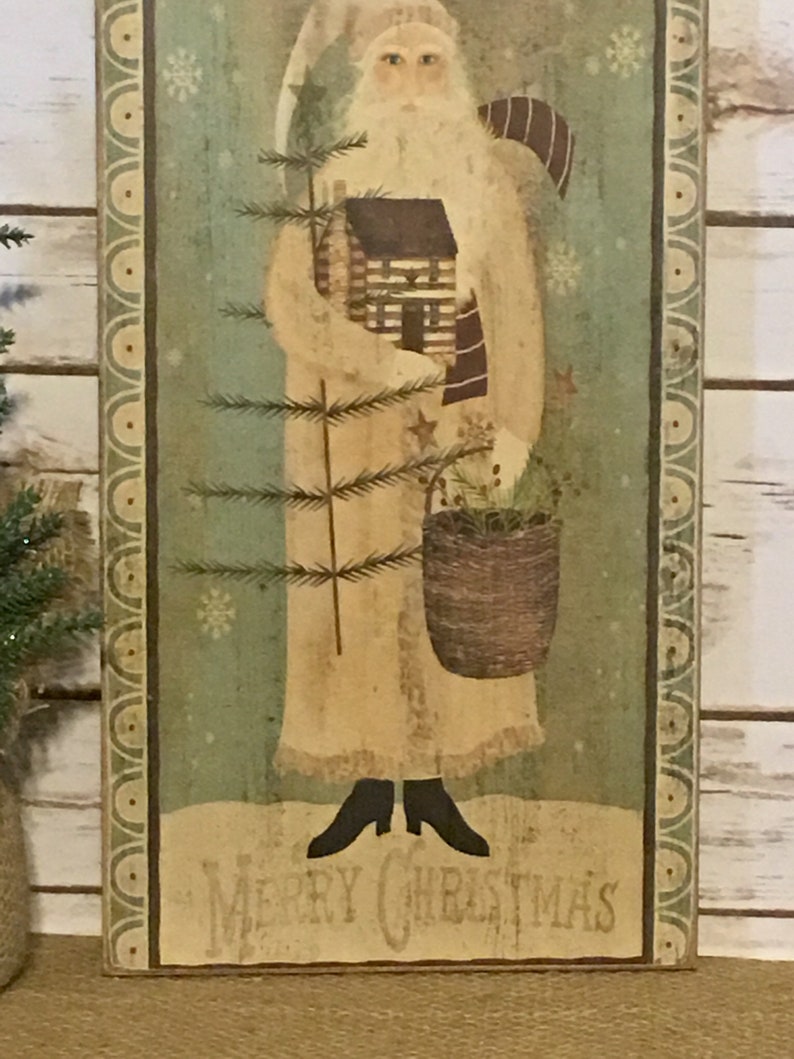 May include: A distressed wooden Christmas sign with a folk art image of Santa Claus. Santa is in a cream robe, holding a house and basket. The sign has a green background with snowflakes and the words "Merry Christmas" at the bottom.
