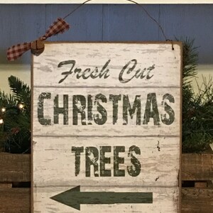 以下が含まれることがあります： 「Fresh Cut CHRISTMAS TREES」の文字が黒字で書かれた長方形の木製看板。矢印は左を指しています。看板にはワイヤーハンガーと、赤と白のチェック柄のリボンが付いています。