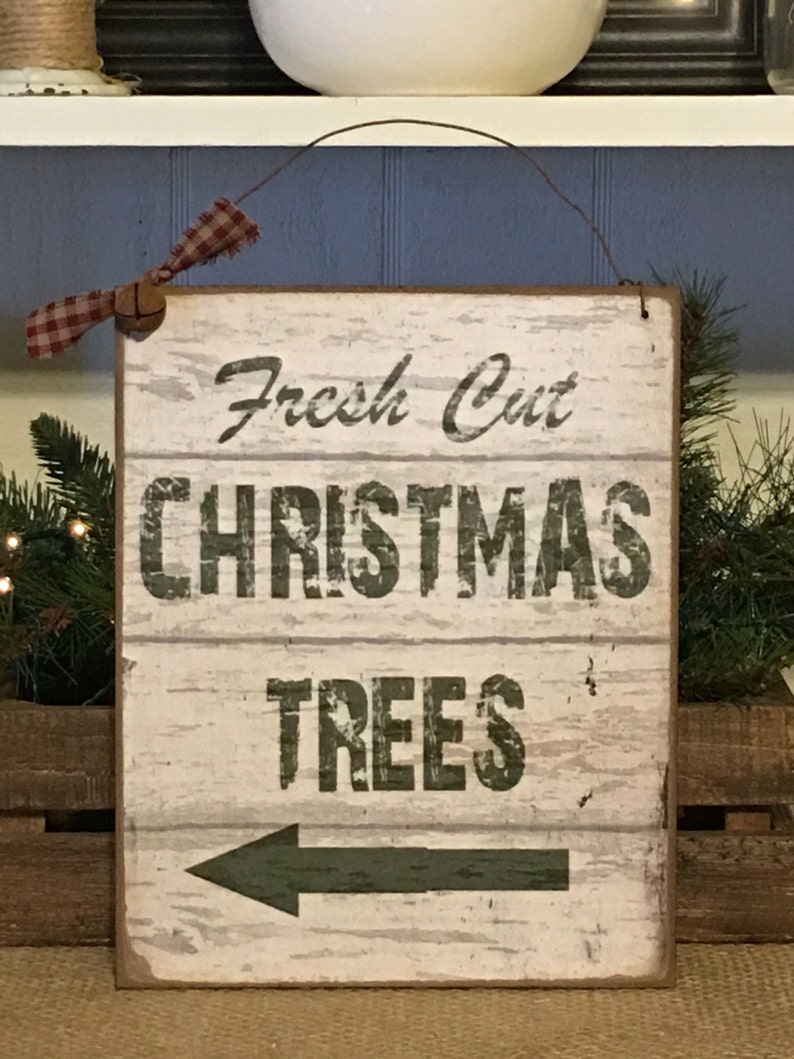 以下が含まれることがあります： 「Fresh Cut CHRISTMAS TREES」の文字が黒字で書かれた長方形の木製看板。矢印は左を指しています。看板にはワイヤーハンガーと、赤と白のチェック柄のリボンが付いています。