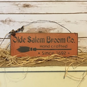 May include: A rectangular, orange wooden sign with the text "Olde Salem Broom Co." in dark brown lettering. Below the text is a black broom graphic and the words "Hand crafted Since 1692." The sign is suspended by a wire and decorated with raffia.