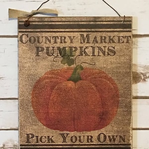 Puede incluir: Un letrero rectangular de madera con el texto "COUNTRY MARKET PUMPKINS PICK YOUR OWN" en letras marrones. El letrero presenta una gran ilustración de calabaza naranja y está decorado con un lazo de arpillera y un colgador de alambre.