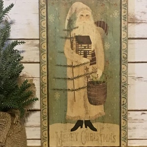 May include: A rectangular wooden sign with a folk art image of Father Christmas. He wears a cream-coloured robe and hat, holding a small cabin and a Christmas tree. The sign has a green background with snowflakes and the words "Merry Christmas".