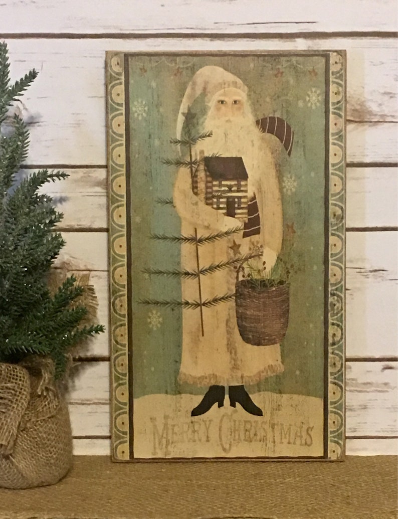 May include: A rectangular wooden sign with a folk art image of Father Christmas. He wears a cream-coloured robe and hat, holding a small cabin and a Christmas tree. The sign has a green background with snowflakes and the words "Merry Christmas".