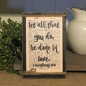 May include: A wooden sign with a black background and a distressed white finish. The sign features a bible verse from 1 Corinthians 16:14, "Let all that you do, be done in love." The text is printed in black on a vintage dictionary page.