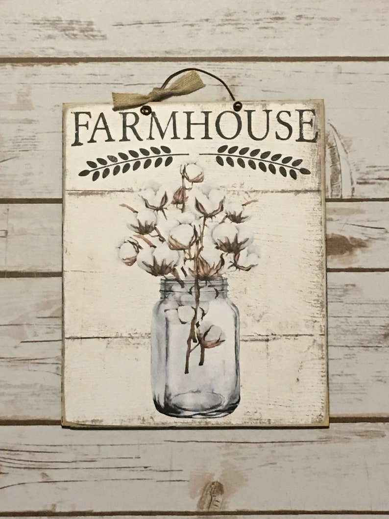 May include: White wooden sign with the text "FARMHOUSE" in black lettering with a decorative wreath above and below the text. The sign features a painted image of a glass jar filled with cotton bolls.