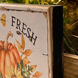 May include: A rustic wooden sign with the word "FRESH" in black lettering. The sign features a watercolor illustration of a pumpkin, sunflowers, and fall leaves in shades of orange, yellow, and brown. The edges of the sign are distressed.